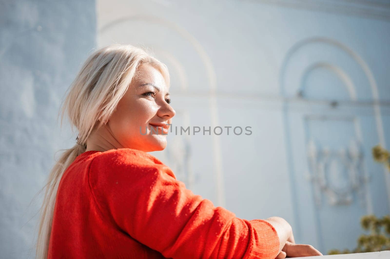 A woman in a red sweater is sitting on a ledge. She is smiling and looking out at the street