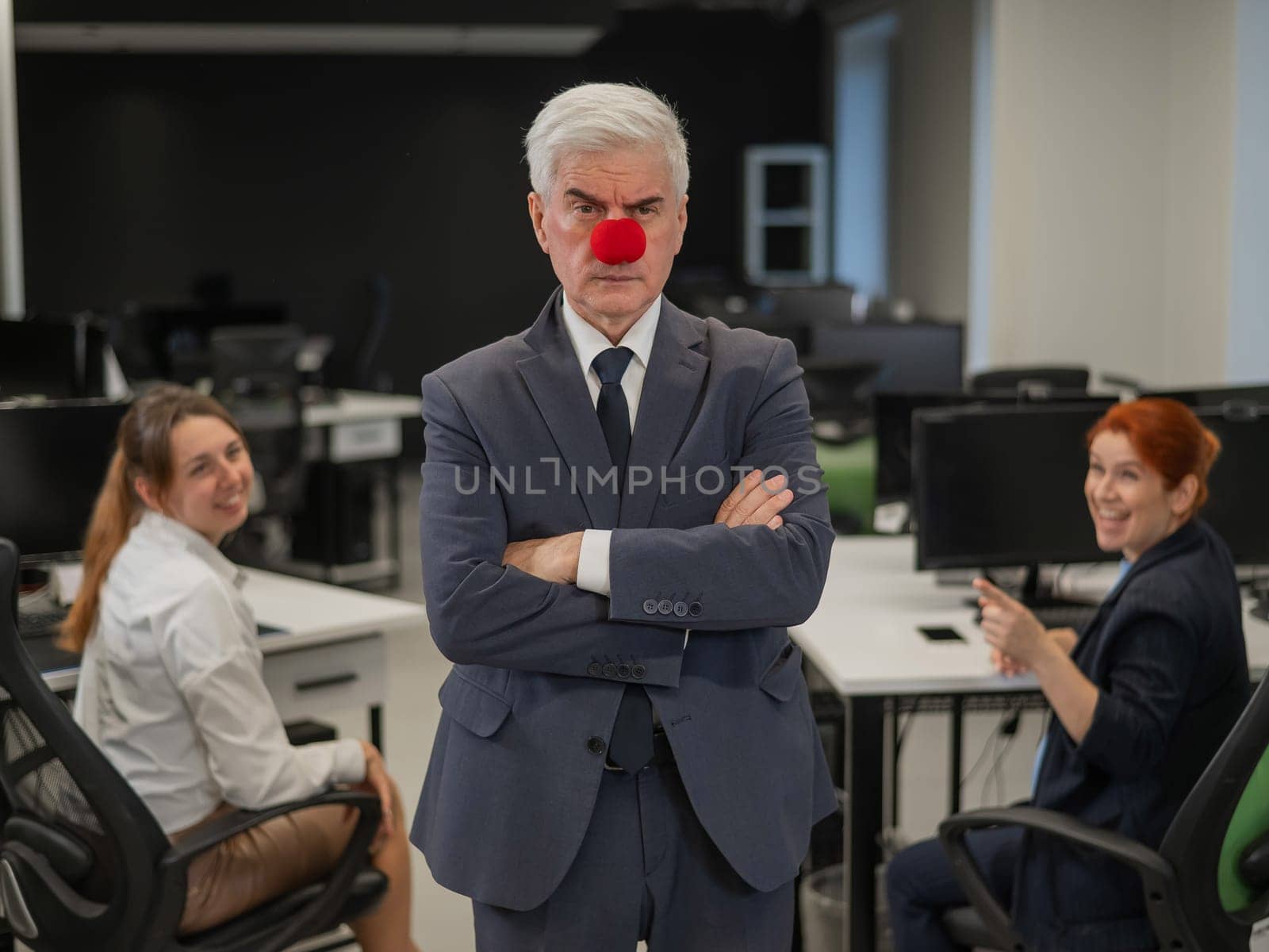 Two Caucasian women look at a serious elderly man with a clown nose in the office