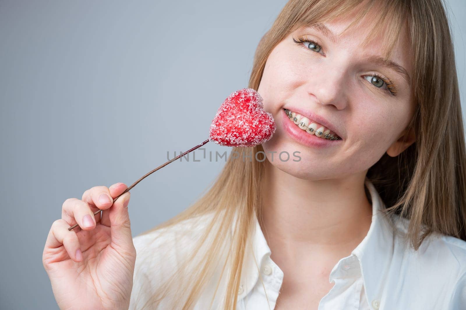 Cute woman with braces on her teeth holds a candy in the form of a heart on white background. Copy space.