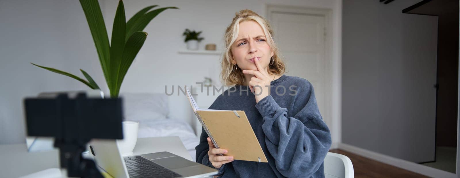 Portrait of young woman, student sitting in room, studying from home, remote education concept, holding notebook and thinking, looking aside with thoughtful face, concentrating on assignment by Benzoix