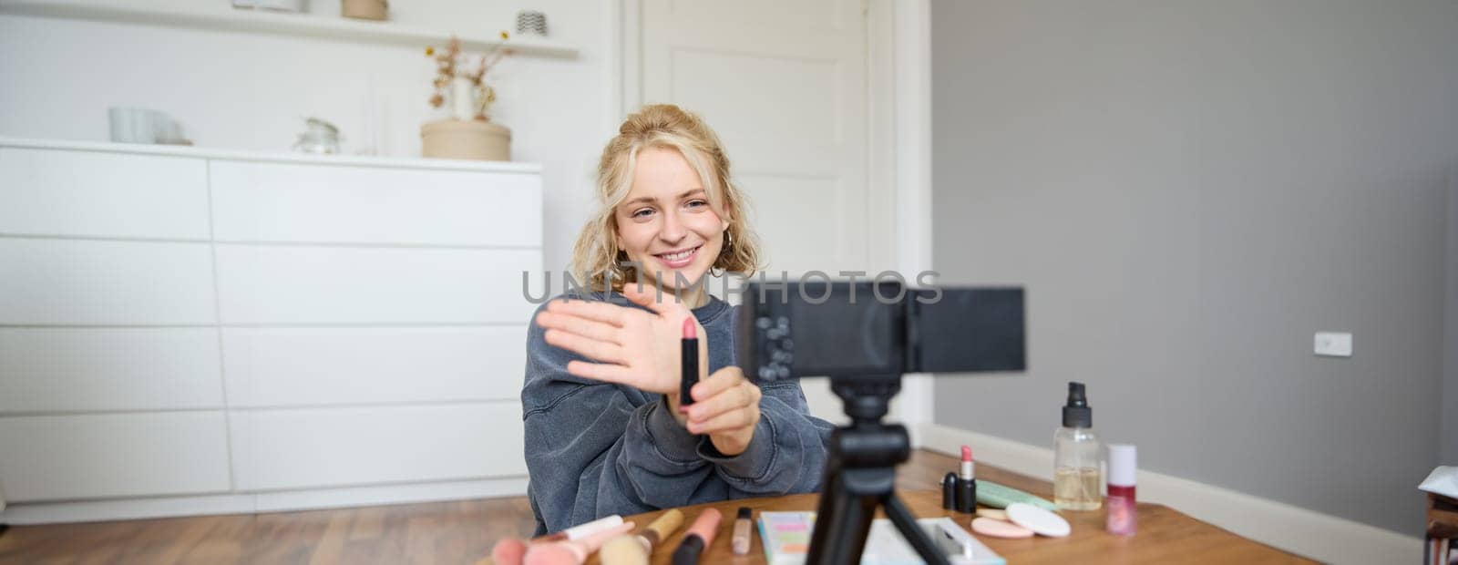 Image of young female content creator, beauty blogger sits on floor in her room, records video about makeup, shows lipstick on camera by Benzoix