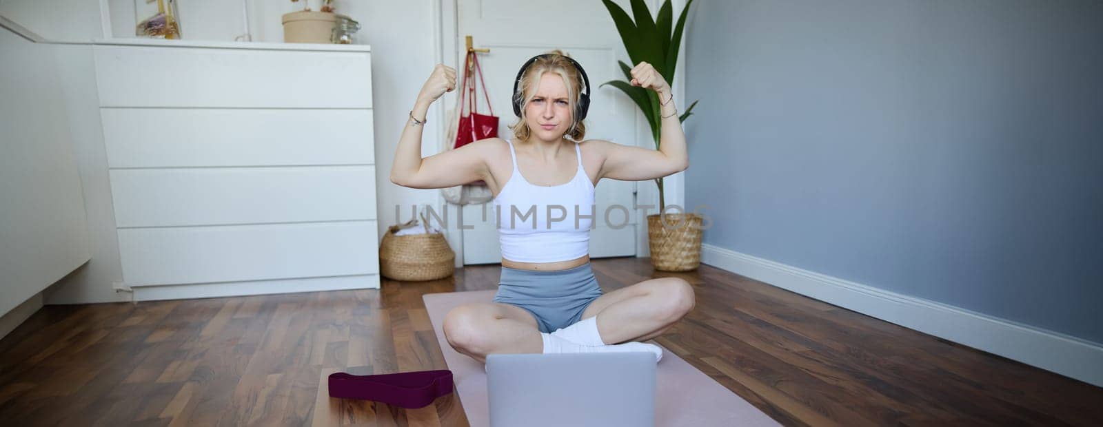 Portrait of young athletic woman doing workout at home, shows her muscles, strong biceps, sits on yoga mat and wears wireless headphones.