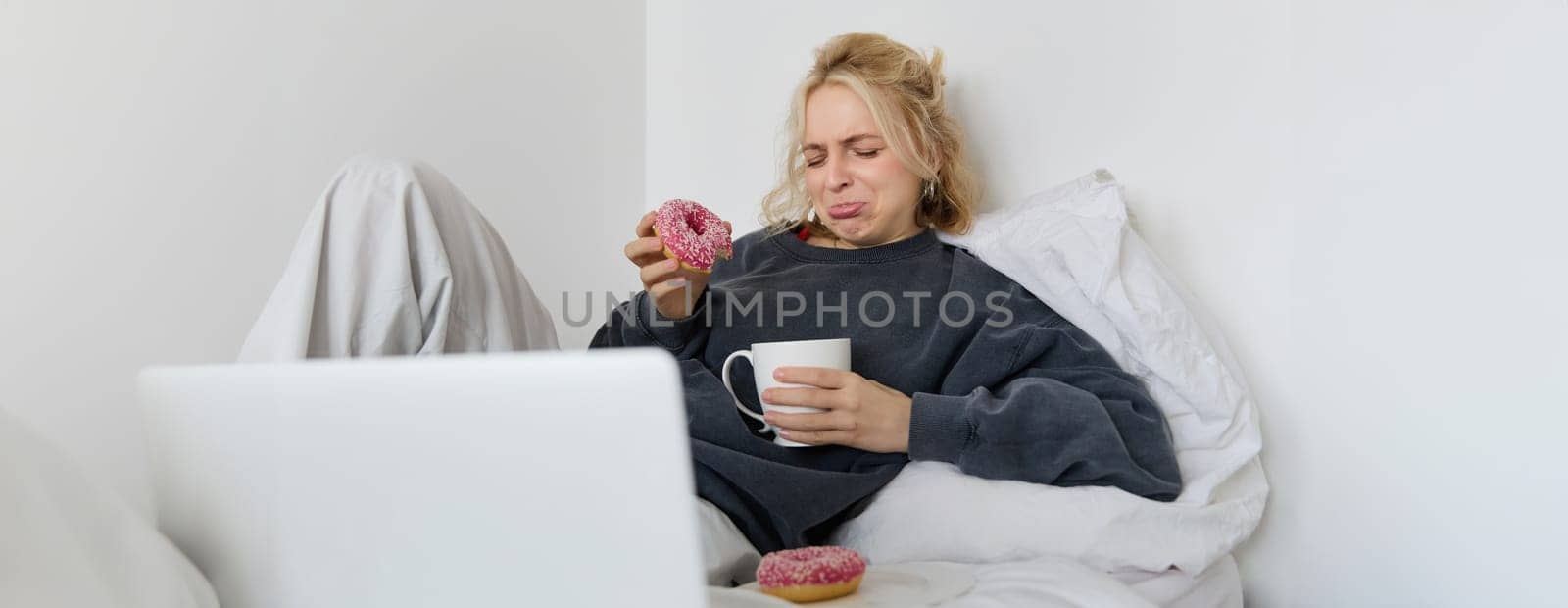Portrait of woman watching sad movie on laptop, crying and wiping tears off, eating doughnut and drinking tea.