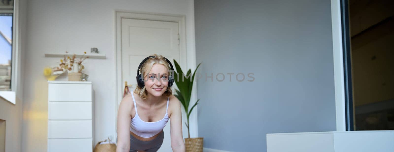 Vertical shot of young blond woman at home, wearing wireless headphones, looking at laptop, standing in plank, watching fitness tutorial online.