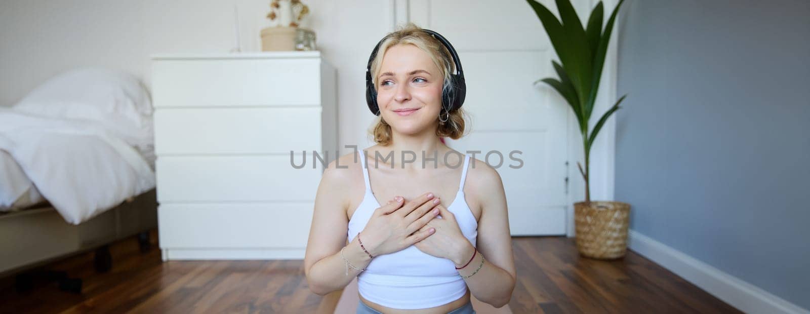 Portrait of young blond woman in headphones, feeling calm and relaxed after meditation, yoga training, sitting on yoga mat in her room. Wellbeing, sport and people concept