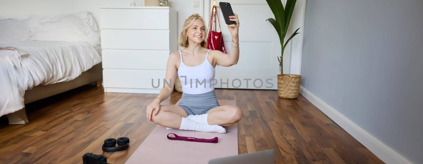 Portrait of stylish young woman, sitting on rubber mat on floor at home, taking selfie while doing workout, yoga session, posting photo on social media by Benzoix