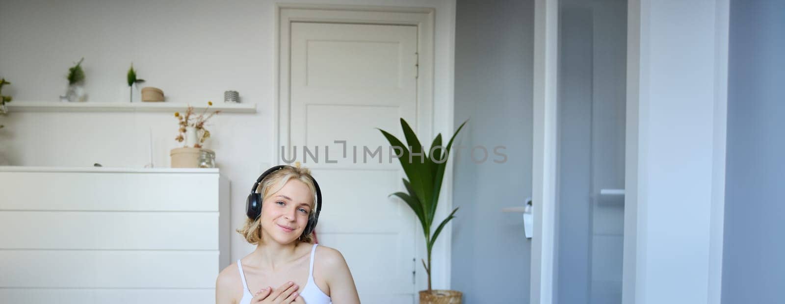 Portrait of young blond woman in headphones, feeling calm and relaxed after meditation, yoga training, sitting on yoga mat in her room by Benzoix