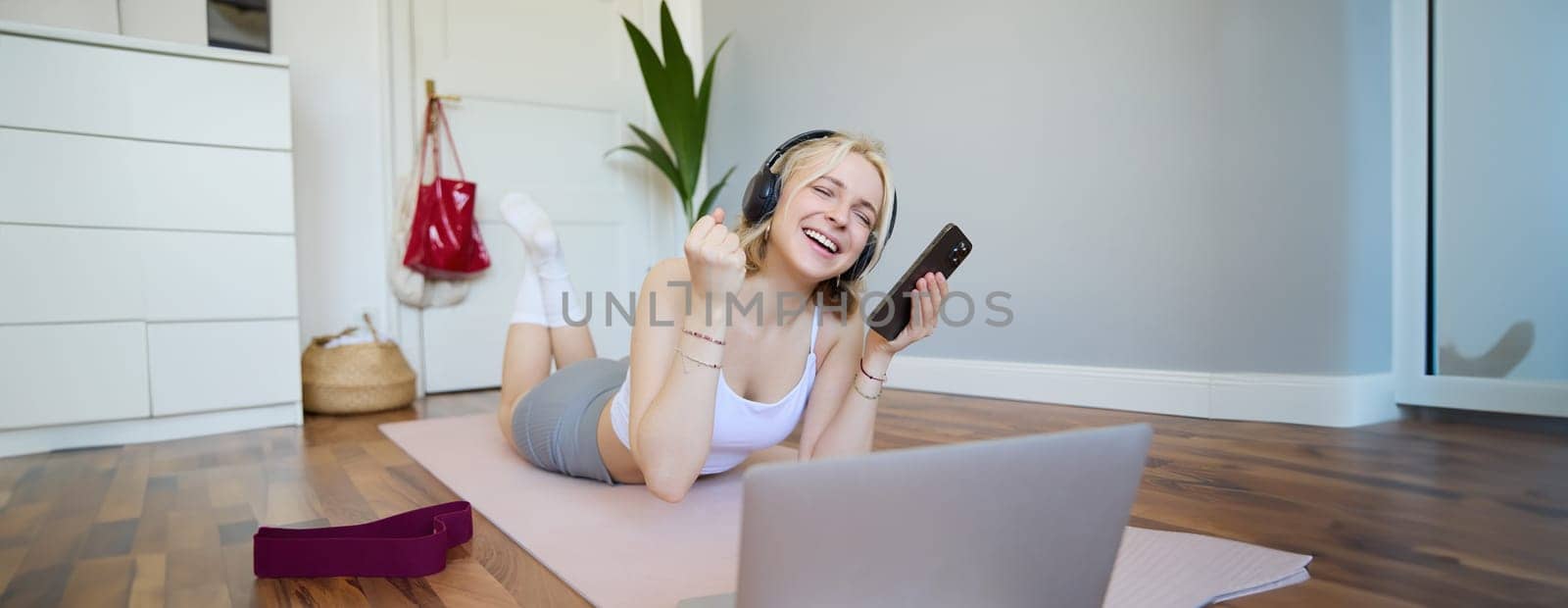 Young woman doing fitness workout at home, lying on rubber yoga mat, holding smartphone, wearing headphones, using laptop to watch sports videos by Benzoix