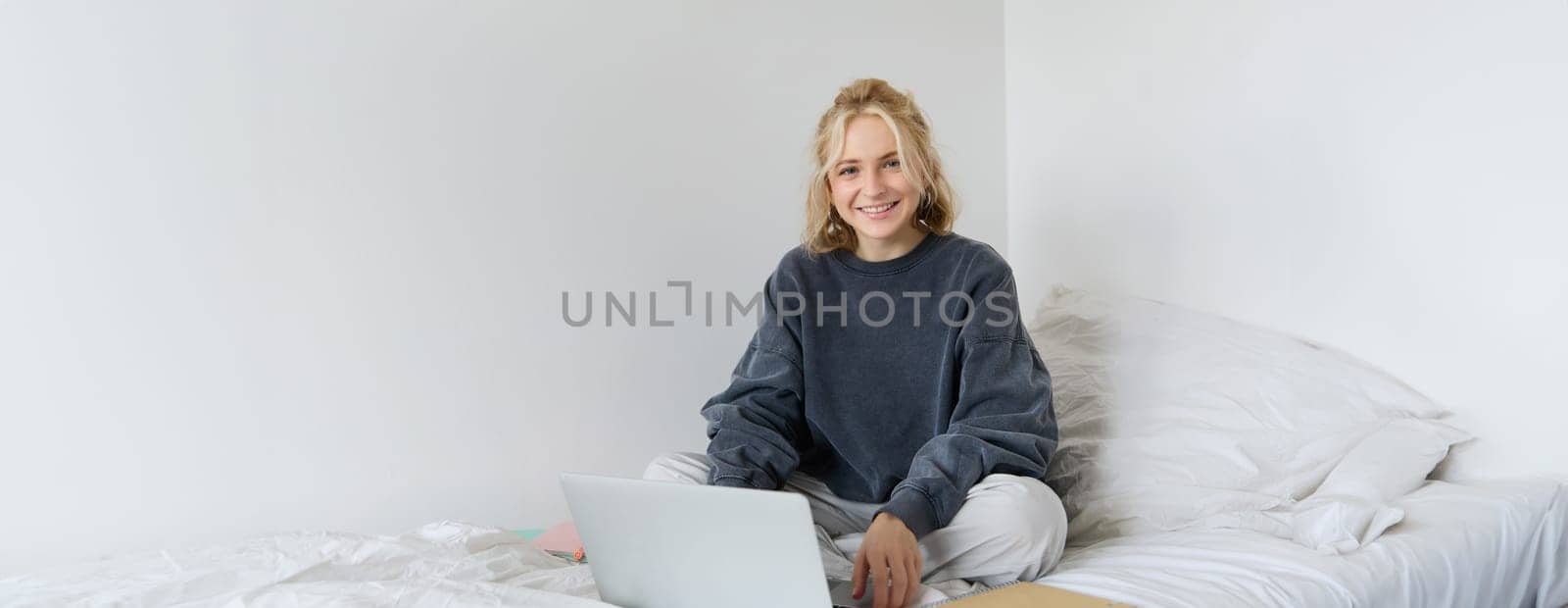 Portrait of young smiling woman studying in her bed, working from home in bedroom, sitting with laptop and notebooks on lotus pose, looking happy and relaxed by Benzoix