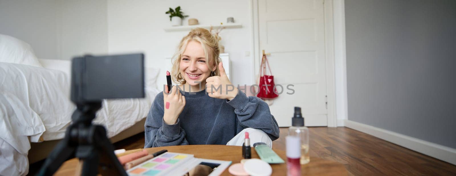 Happy young content creator, woman showing lipstick, recording lifestyle, beauty vlog on digital camera, makes thumbs up hand gesture, recommending makeup product.
