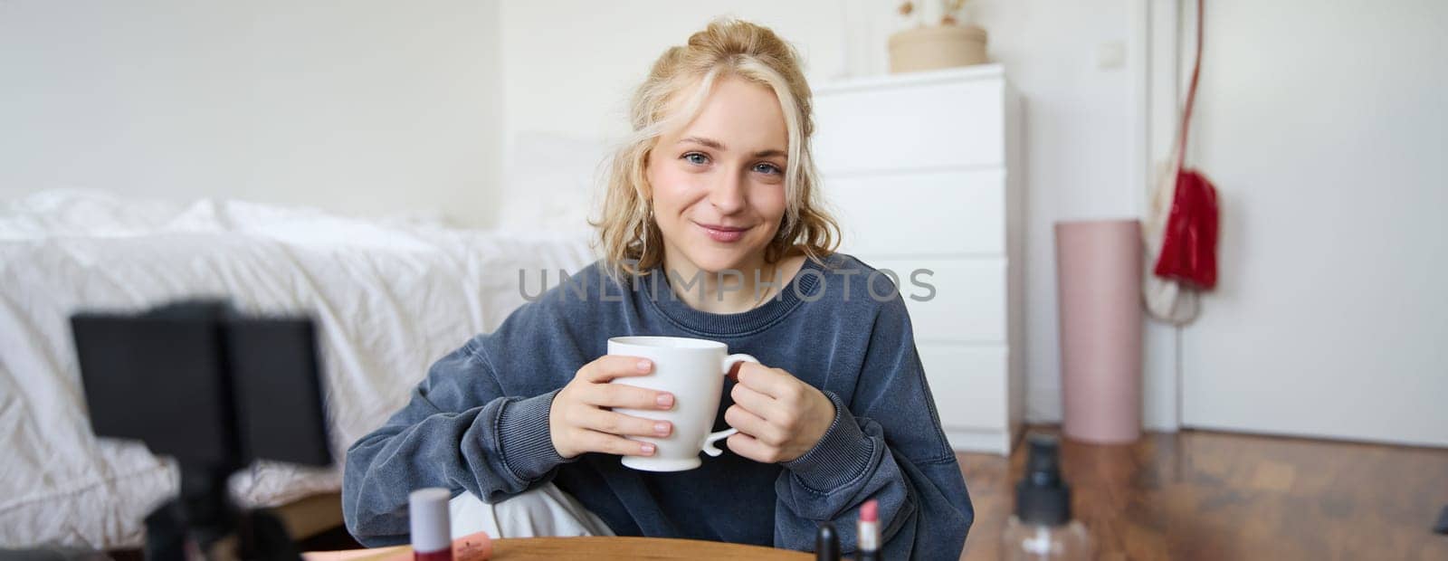Close up portrait of beautiful blond woman, girl sits in her room, does makeup, records video for social media, drinks tea, holds mug and smiles at camera by Benzoix