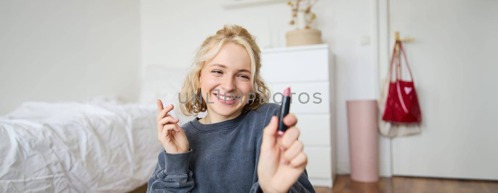 Portrait of young social media influencer, woman recording a video with beauty products, showing her makeup on camera, holding lipstick and smiling, sitting on floor by Benzoix