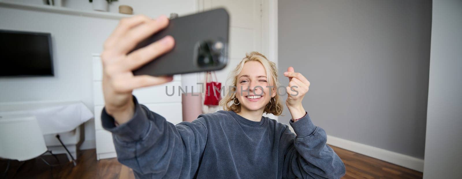 Portrait of young stylish girl sits on bedroom floor, takes selfies on her smartphone, posing for photo on social media app, smiling and looking happy at camera by Benzoix