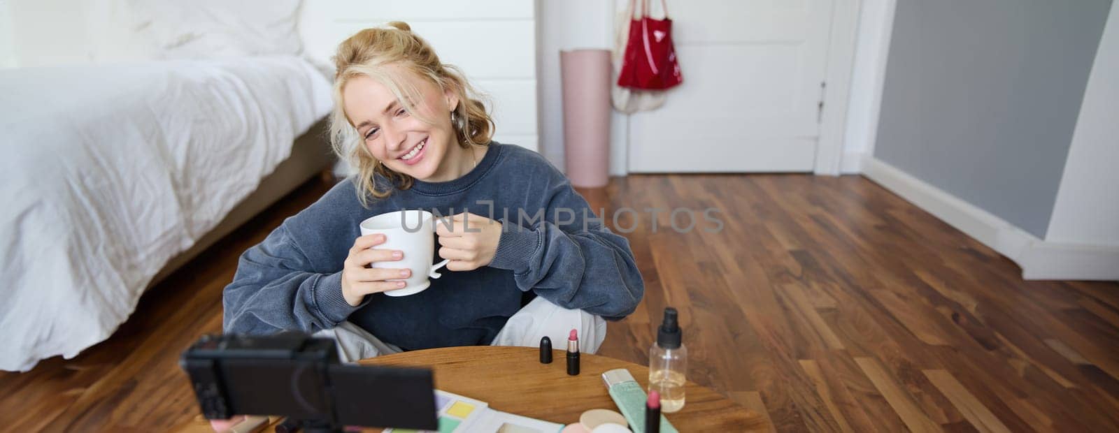 Young woman smiling, recording lifestyle vlog on her digital camera, holding cup of tea while talking, has makeup on coffee table, doing makeup tutorial for social media from her room by Benzoix