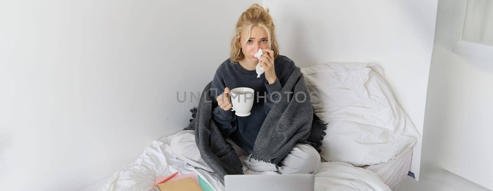 Portrait of young woman sitting at home sick, feeling unwell, drinking tea, spending time in bed, has runny nose, working on laptop by Benzoix
