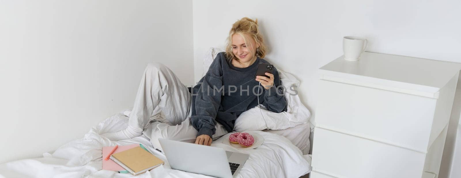 Lifestyle and weekend concept. Young smiling woman, relaxing at home, lying in bed with cup of tea and doughnut, using laptop and smartphone in bedroom.