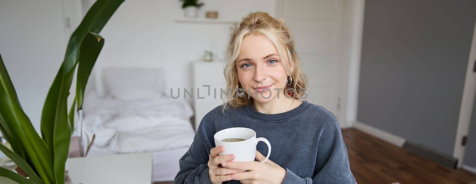 Portrait of smiling beautiful young woman, sitting in bedroom with cup of tea, resting at home alone, enjoys her weekend indoors.