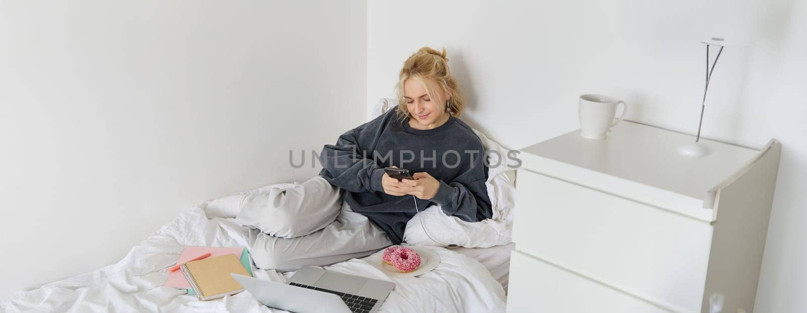 Lifestyle and people concept. Young beautiful woman, staying at home, lying in bed with laptop and smartphone, eating doughnut, enjoying free time, spending weekend at home, watching movie online.