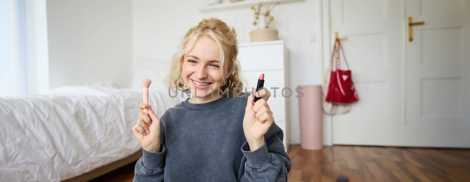 Image of young female content creator, beauty blogger sits on floor in her room, records video about makeup, shows lipstick on camera.