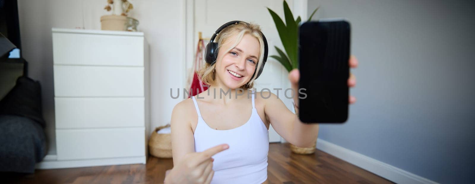 Portrait of smiling, fit and healthy, fitness instructor at home, pointing finger and showing her smartphone screen, recommending sports app for workout.