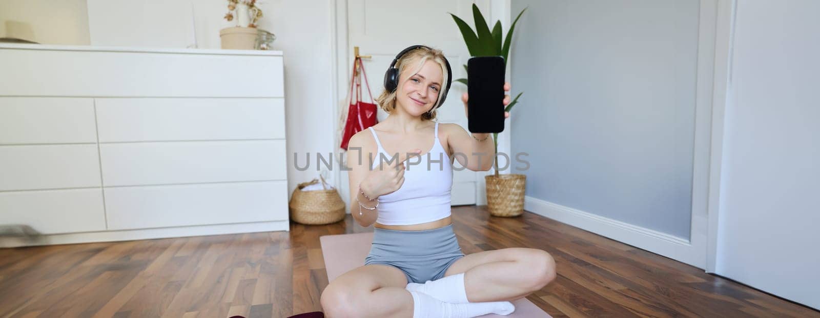 Portrait of fitness instructor showing her favourite workout app, pointing finger at smartphone screen, showing mobile app on camera, smiling and looking satisfied, wearing headphones.