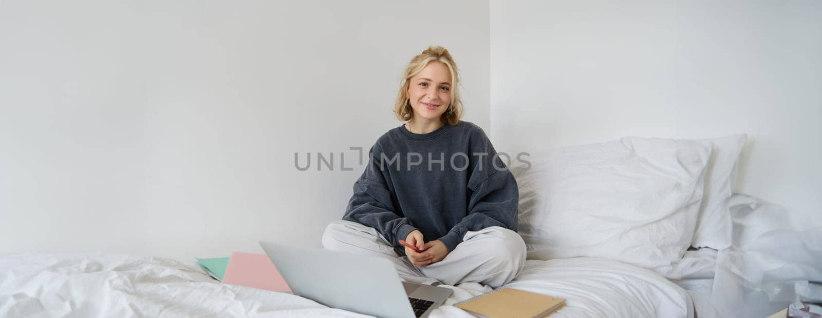 Portrait of female student, woman sits on bed with laptop and notebooks, studying online, remote education concept. Girl freelancer working from her bedroom.