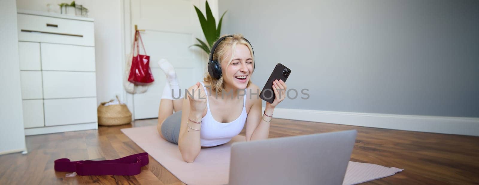 Portrait of young woman workout, watching exercise videos on laptop in headphones, lying on rubber mat with mobile phone and smiling by Benzoix