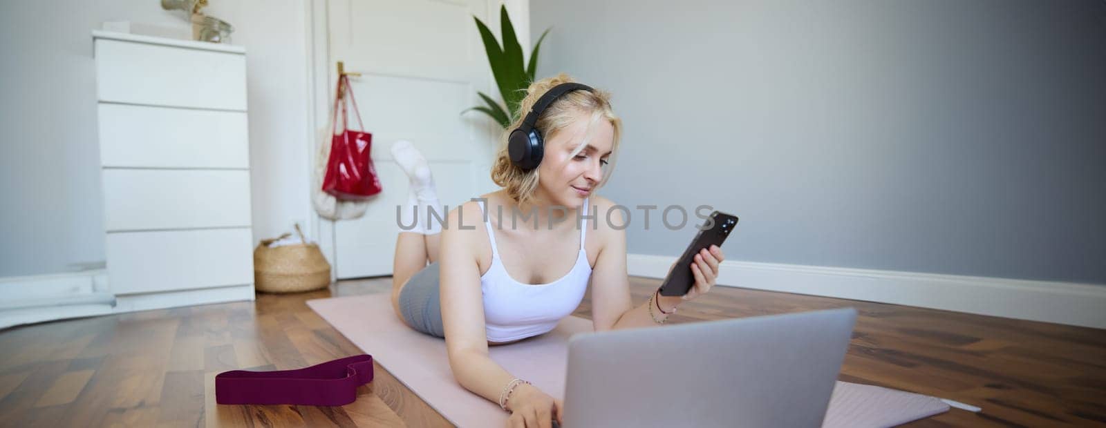 Portrait of happy, smiling sporty woman, doing workout at home, using laptop to watch workout videos in headphones, checking sports app on mobile phone by Benzoix