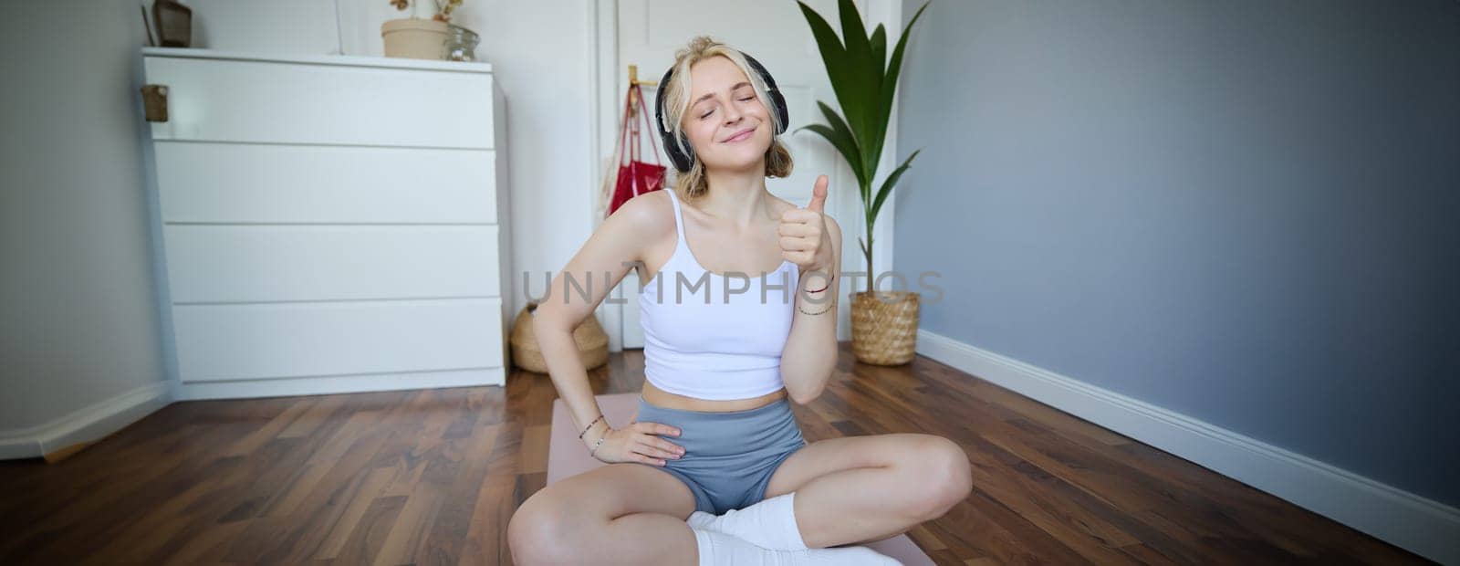 Portrait of happy, fit and healthy young woman sitting on yoga mat, wearing wireless headphones, showing thumbs up with satisfied face, pleased with workout training at home by Benzoix