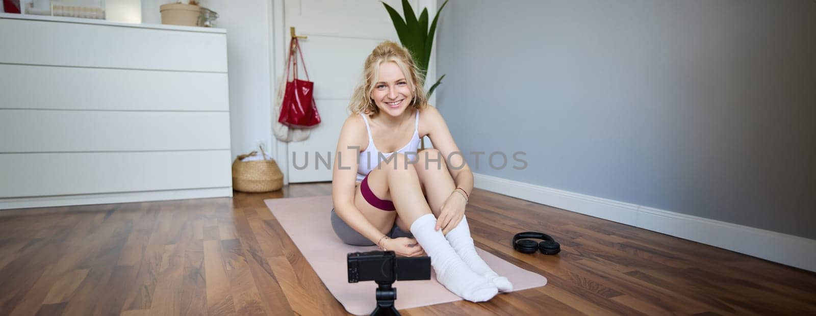 Portrait of young blond woman, social media vlogger using digital camera during workout, shooting video about exercises and fitness, stretching with rubber resistance band.