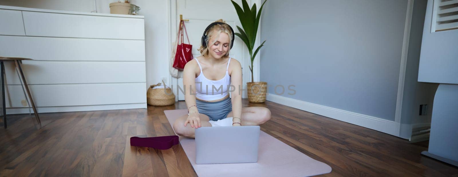 Portrait of woman during workout, sitting on yoga mat with resistance band, listening to video instructions on laptop, wearing wireless headphones, repeating exercises.