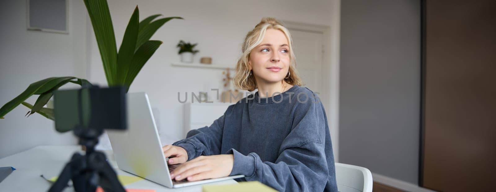 Portrait of smiling young woman, college student sits in her room, does homework, studies remotely from home, uses laptop for freelance work by Benzoix