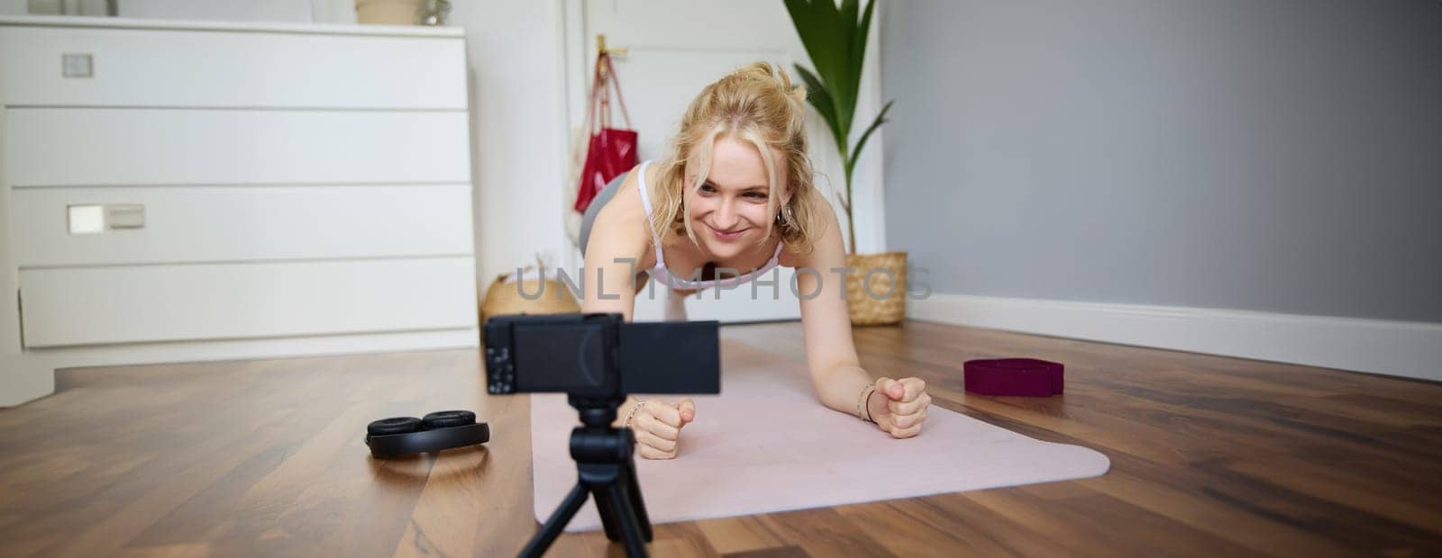 Portrait of young sporty vlogger, fitness instructor standing in plank on rubber yoga mat, recording video of herself doing workout at home by Benzoix