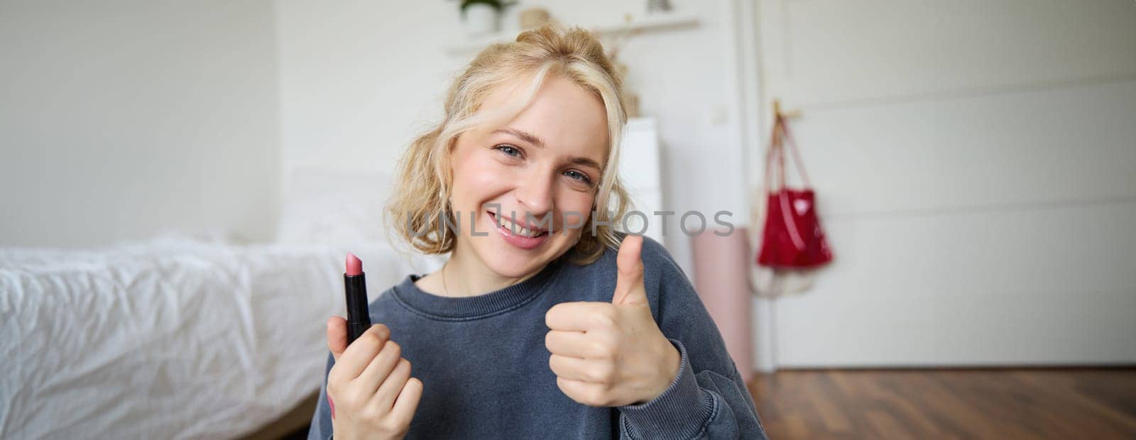 Cute smiling girl shows lipstick and thumbs up hand sign, recommending beauty product, records content for social media account, lifestyle vlog.