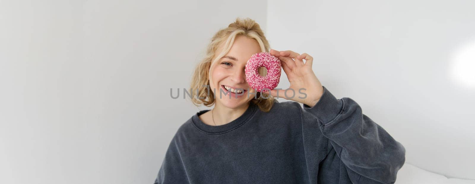 Close up portrait of happy, cute blond woman, holding doughnut, eating sweet, delicious comfort food, showing dessert at camera. Food concept