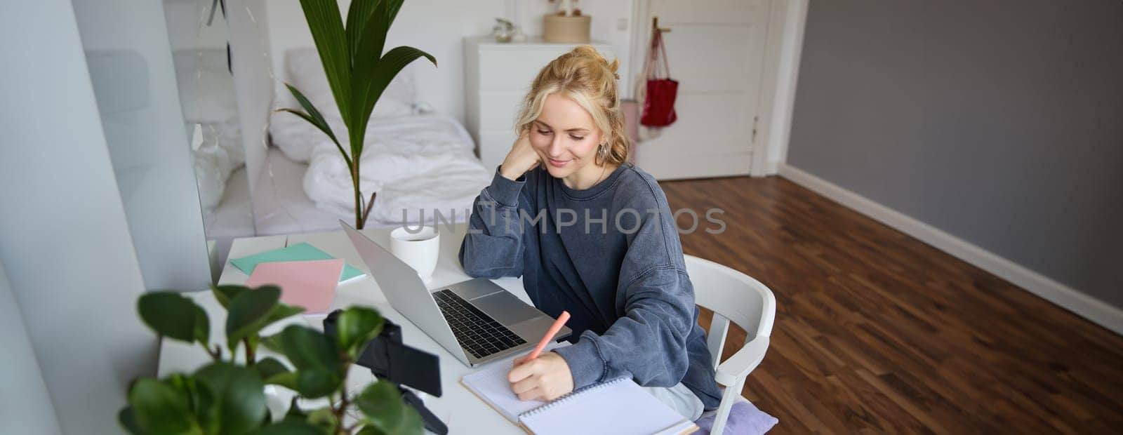 Lifestyle image of young woman writing down something in notebook, making notes, studying online, doing course in internet, listening to interesting information, using laptop by Benzoix