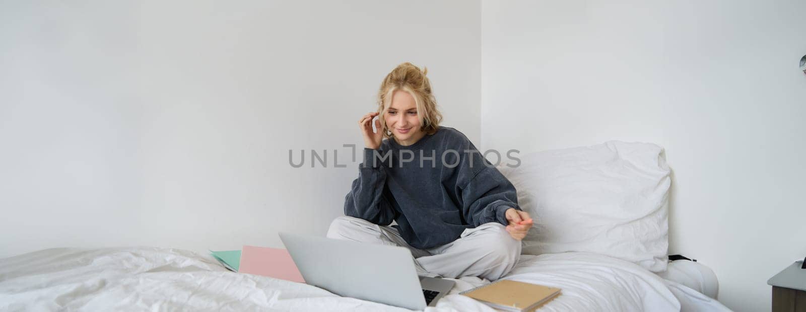 Portrait of female student, woman sits on bed with laptop and notebooks, studying online, remote education concept. Girl freelancer working from her bedroom by Benzoix