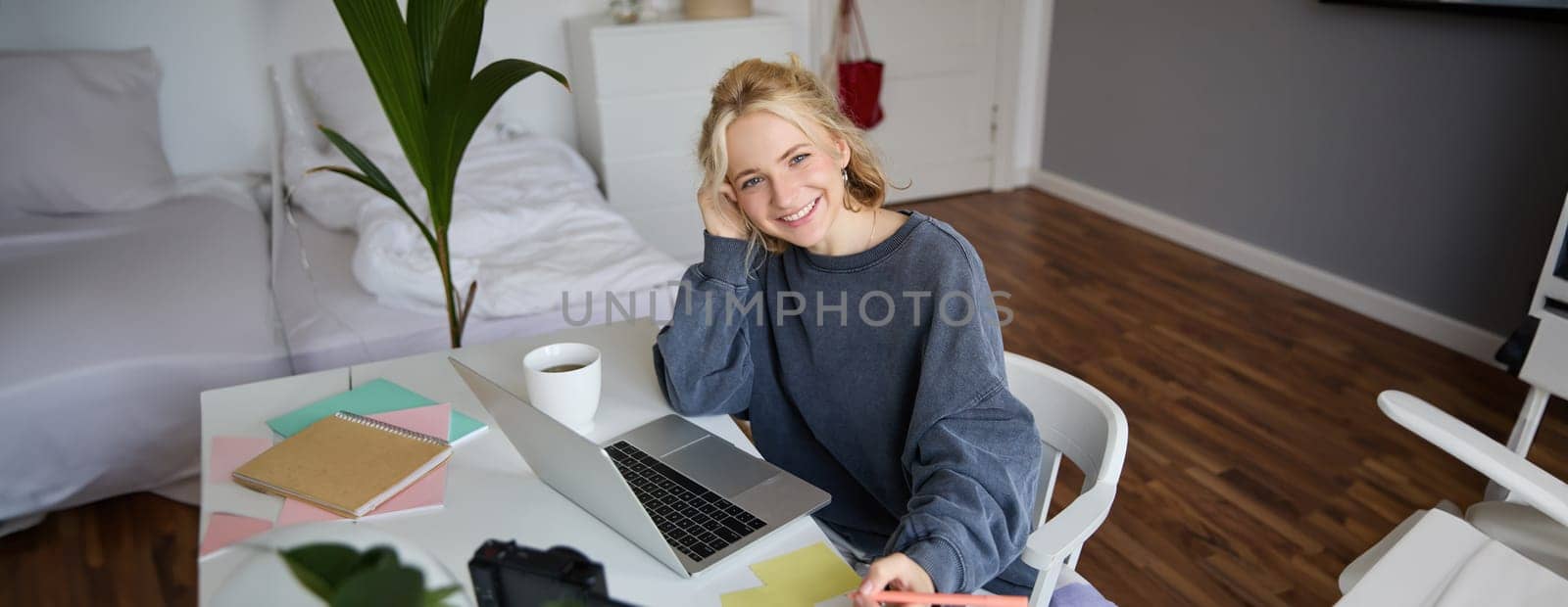 Portrait of smiling, beautiful blond woman, writing down notes, doing homework, studying from home, doing distance learning, online course, working remotely in her room.