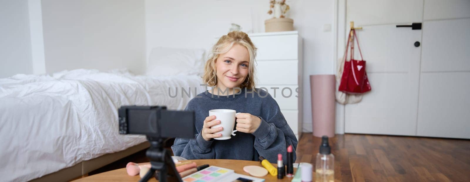 Portrait of beautiful social media beauty blogger, sitting in front of digital camera on floor in bedroom, drinking tea and chatting, talking to followers by Benzoix