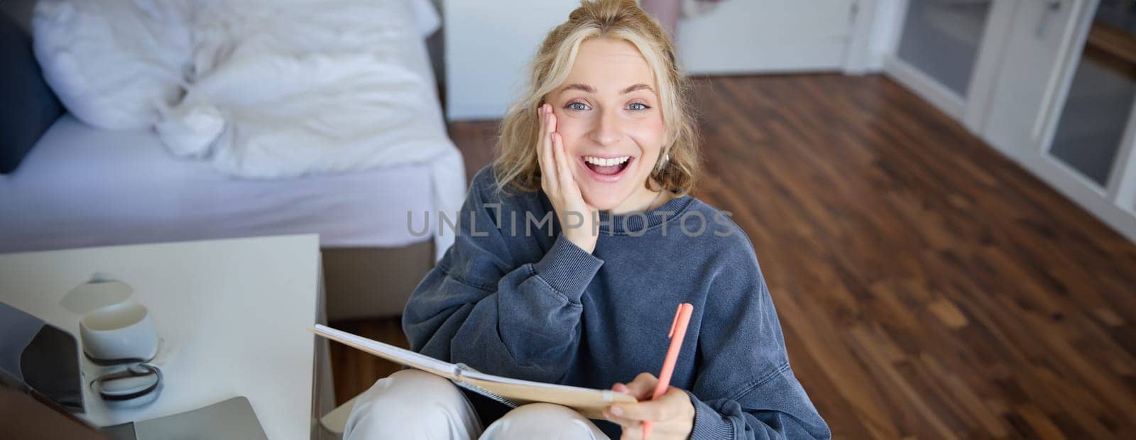 Portrait of happy, young blond woman, sitting in bedroom with notebook and pen, laughing and smiling, writing in journal, doing homework and smiling by Benzoix