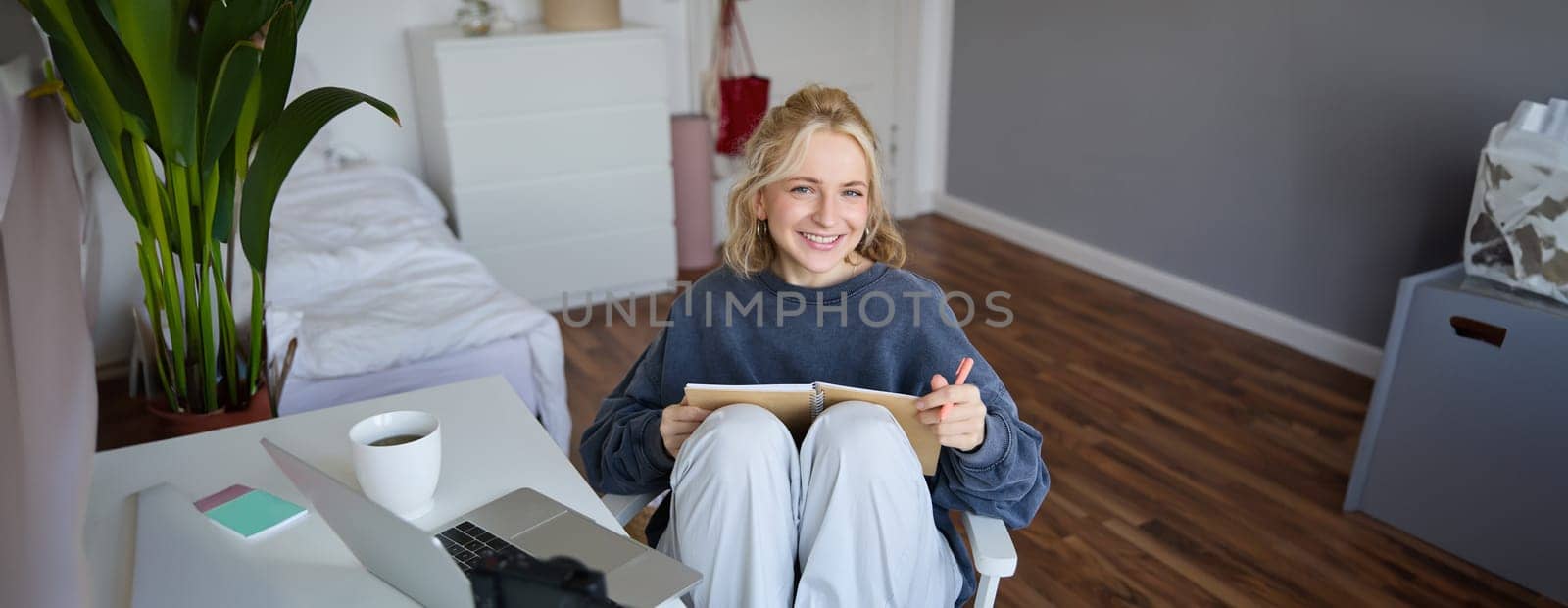 Portrait of young woman reading her notes on journal, sitting at home on chair, smiling and looking happy, doing homework by Benzoix