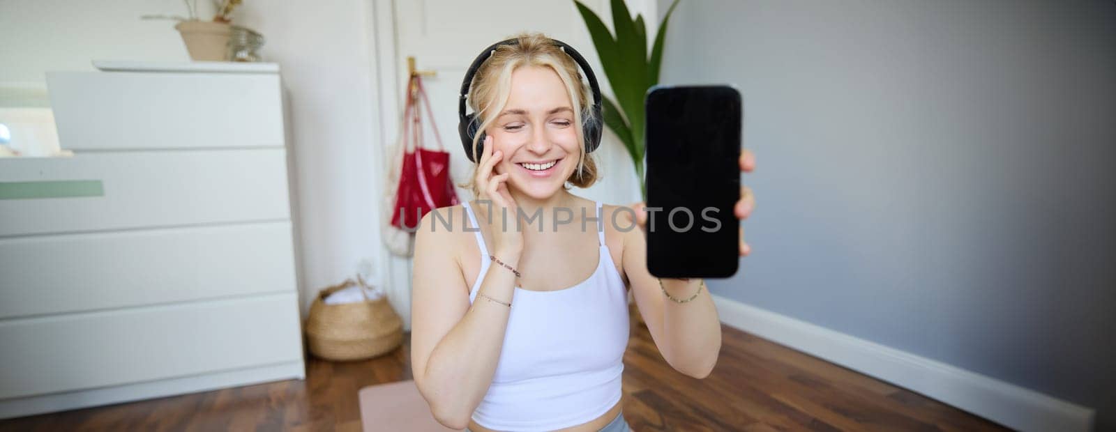 Portrait of smiling, beautiful young woman, showing her smartphone screen, wearing headphones, looking satisfied with sound quality.