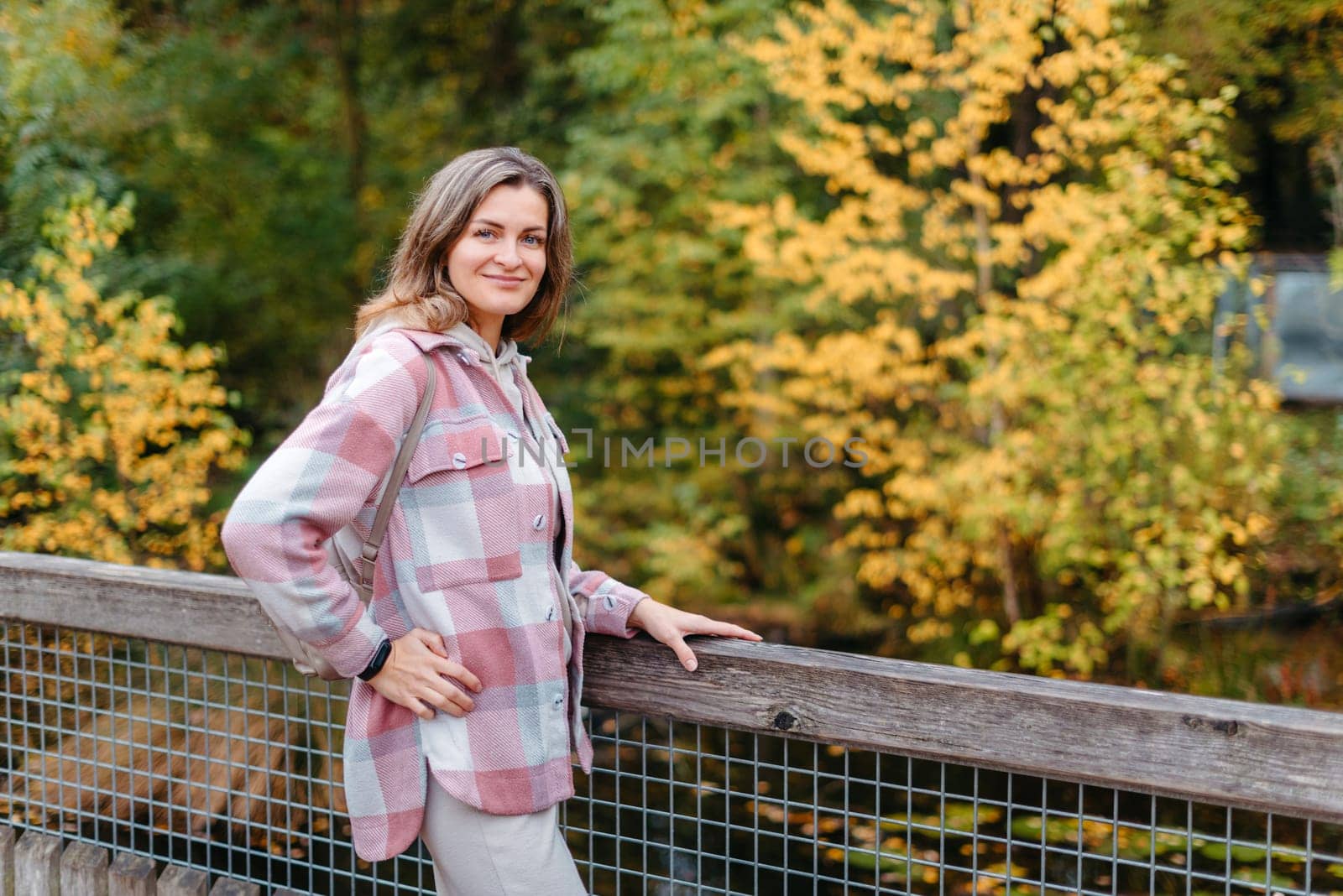 Portrait Of Cute Young Woman In Casual Wear In Autumn, Standing On Bridge Against Background Of An Autumn Park And River. Pretty Female Walking In Park In Golden Fall. Copy Space. Smiling Girl In The Park Standing On Wooden Bridge And Looking At The Camera In Autumn Season by Andrii_Ko