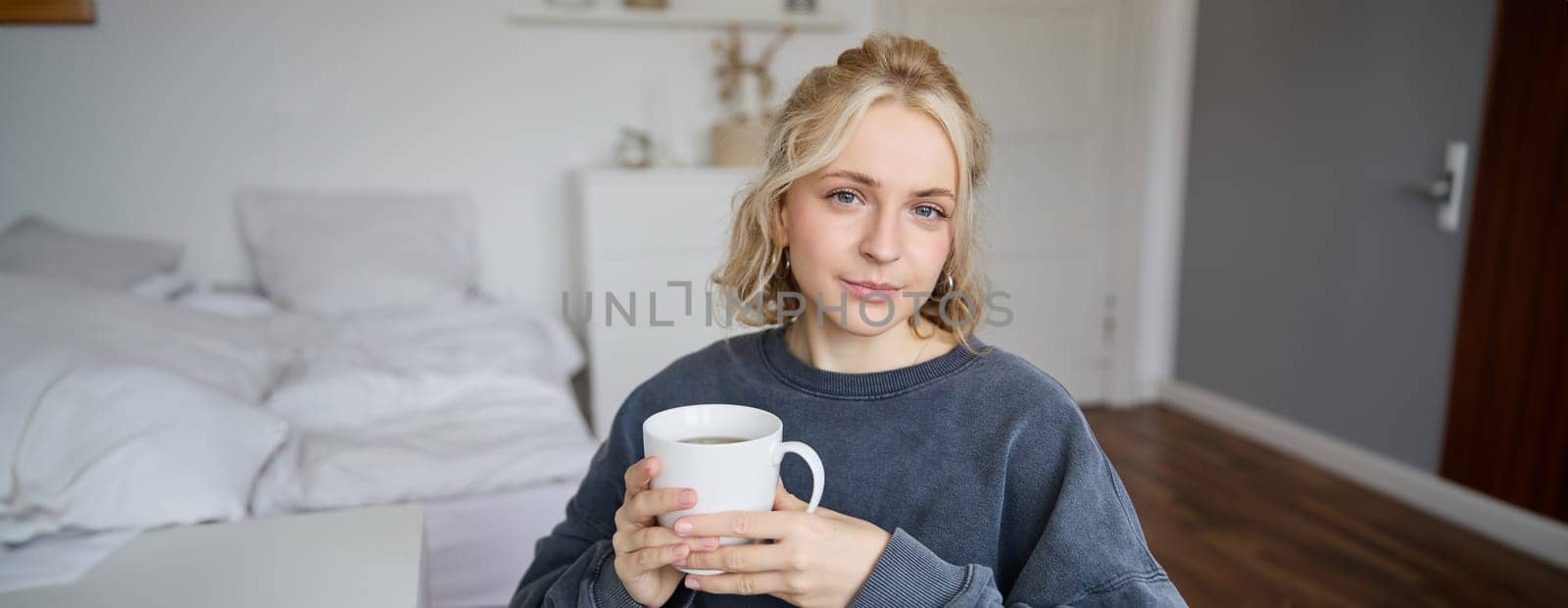 Close up portrait of smiling young blond woman, sits with cup of tea in bedroom, rests at home, enjoys her coffee by Benzoix