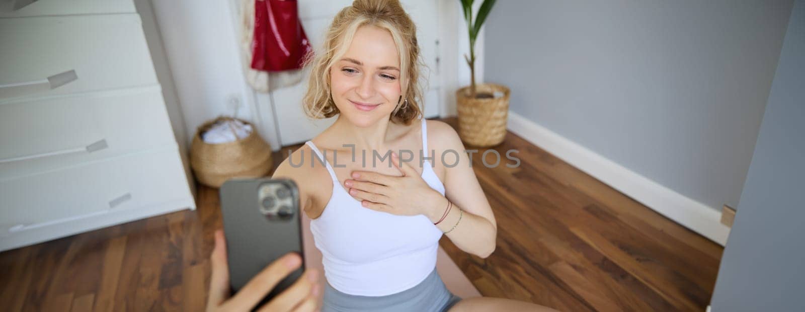 Young sporty fitness trainer, yoga instructor does live stream, takes selfies on smartphone while sitting on rubber mat at home, recording her workout training session by Benzoix