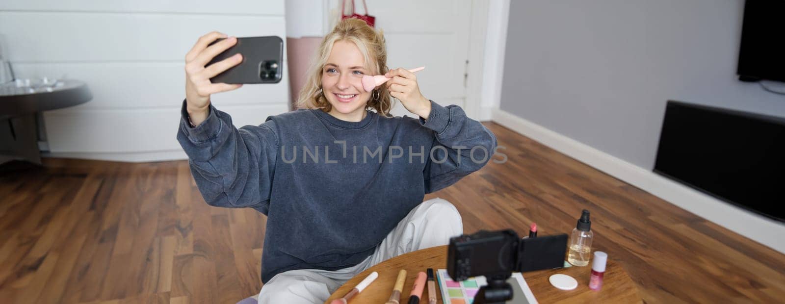Image of stylish young woman, social media influencer, taking pictures on mobile phone, doing makeup tutorial for followers online, recording video vlog in her bedroom, showing brush.