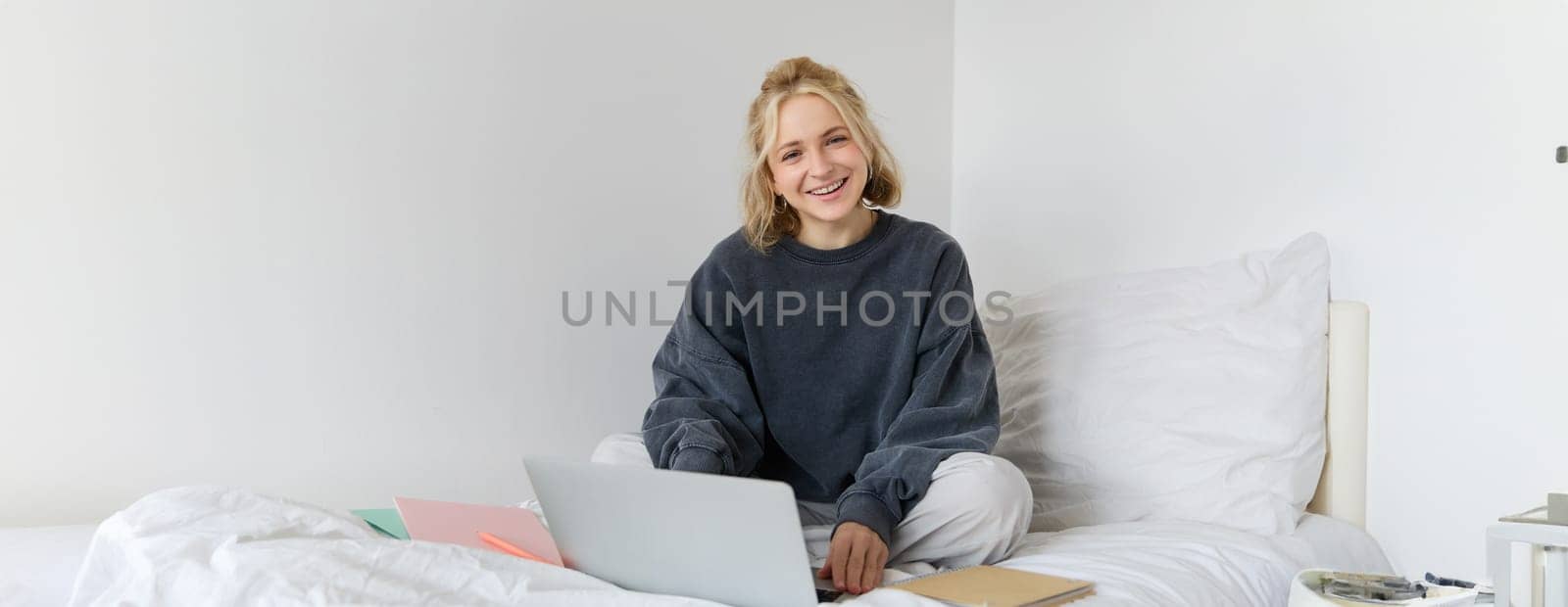 Portrait of young smiling woman studying in her bed, working from home in bedroom, sitting with laptop and notebooks on lotus pose, looking happy and relaxed by Benzoix