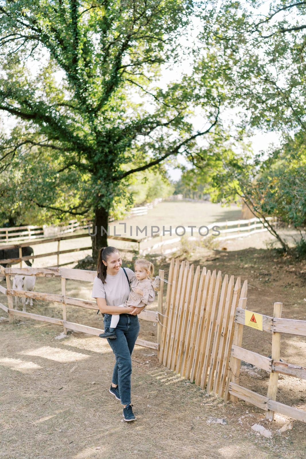 Mother with a little girl in her arms walks past the wooden fence of a paddock with a pony in the park by Nadtochiy