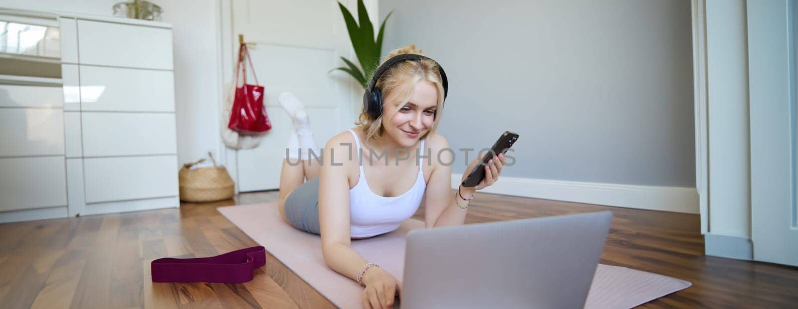 Portrait of young woman workout, watching exercise videos on laptop in headphones, lying on rubber mat with mobile phone and smiling by Benzoix