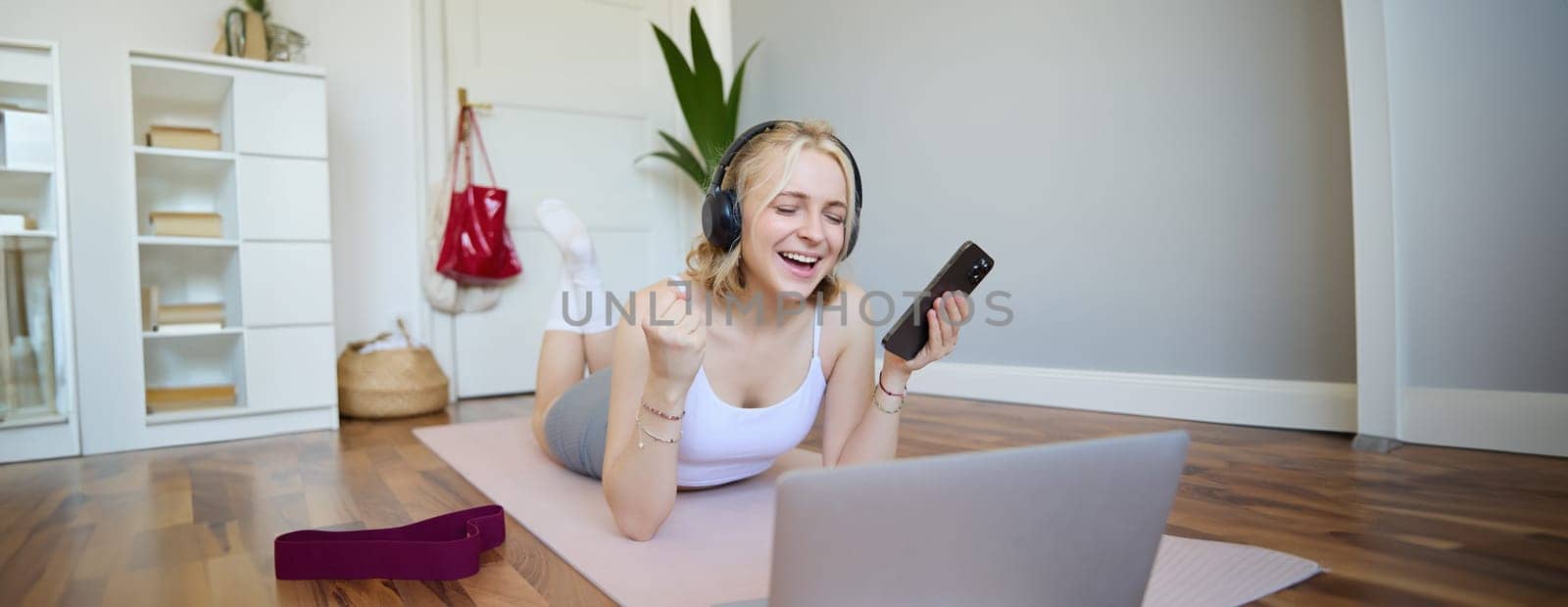 Portrait of happy blond woman in headphones, lying on rubber mat, listens to music in headphones after workout training session, using laptop.